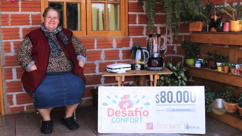 Carmen Chuquiel, ganadora de Desafío Confort, sentada frente a su casa muestra sonriente su cheque y electrodomésticos del premio.