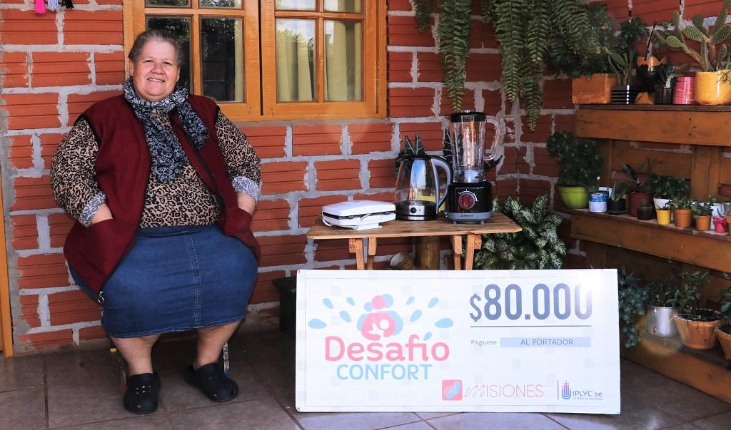 Carmen Chuquiel, ganadora de Desafío Confort, sentada frente a su casa muestra sonriente su cheque y electrodomésticos del premio.