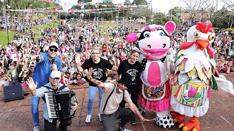 Equipo de Gurises Felices sobre el escenario de la plaza con los niños saludando al fondo, festejando el mes de la infancia.