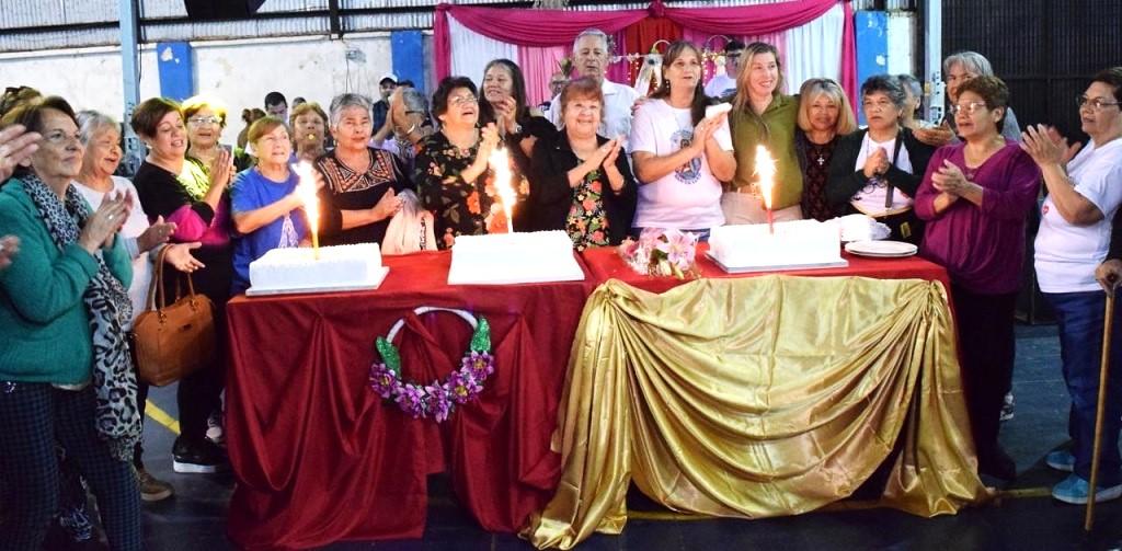 Abuelos cantan feliz cumpleaños alrededor de una mesa con tres tortas con velas encendidas.