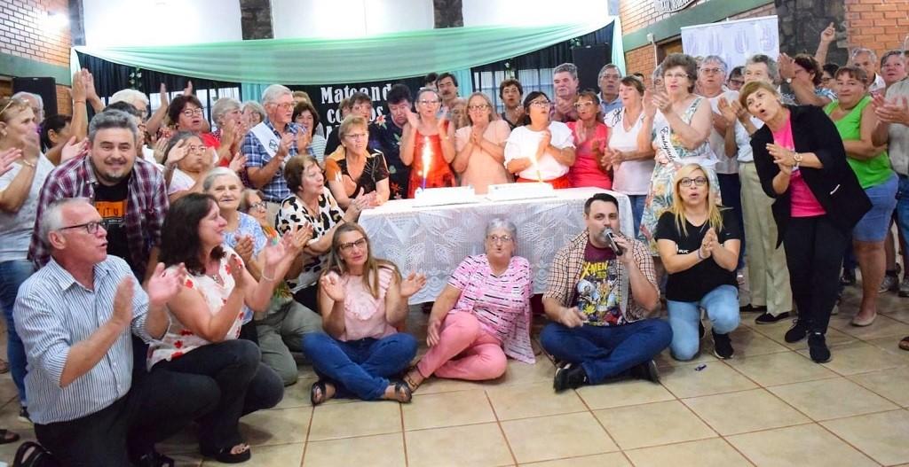 Fiesta de abuelos alrededor de una mesa con tortas cantan y hacen palmas.