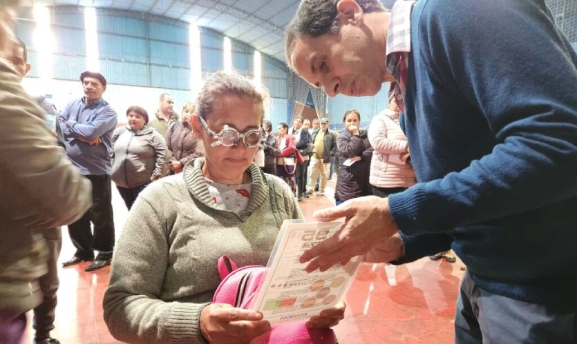 Señora con lentes de prueba óptica lee un texto, asistida por oftalmólogo. Operativo Mirar Mejor en San Javier.