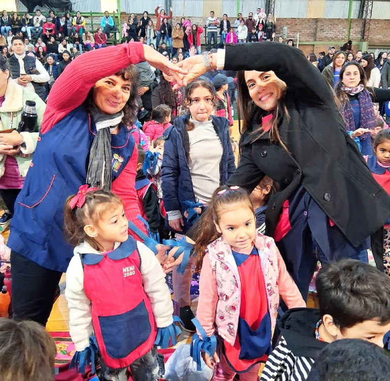 Dos maestras de Jardín hacen un corazón con sus brazos junto a los niños.